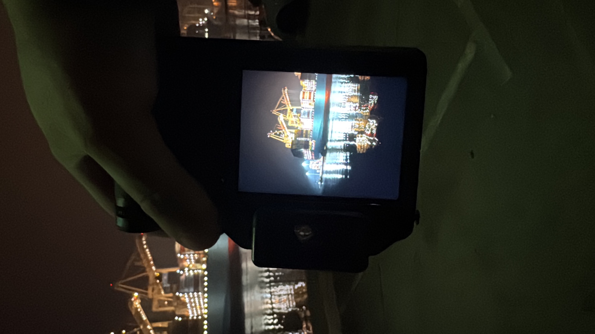 Hands holding a camera while photographing an illuminated container ship at night